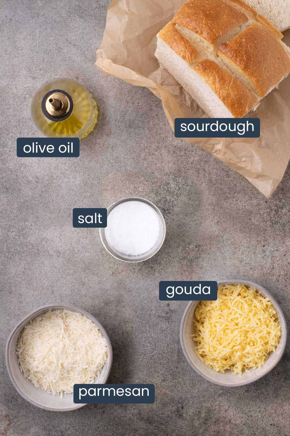 Labeled sourdough bread, olive oil, salt, grated Gouda, and Parmesan arranged on a gray surface.
