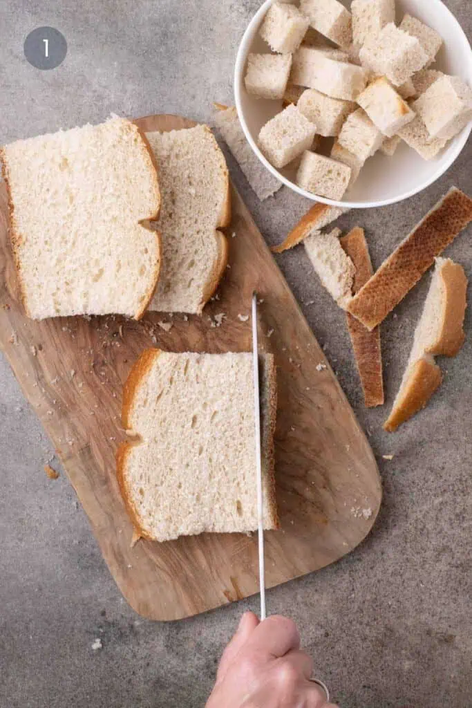 Removing the crust from a sourdough slice beside a bowl of bread cubes and trimmed crusts.