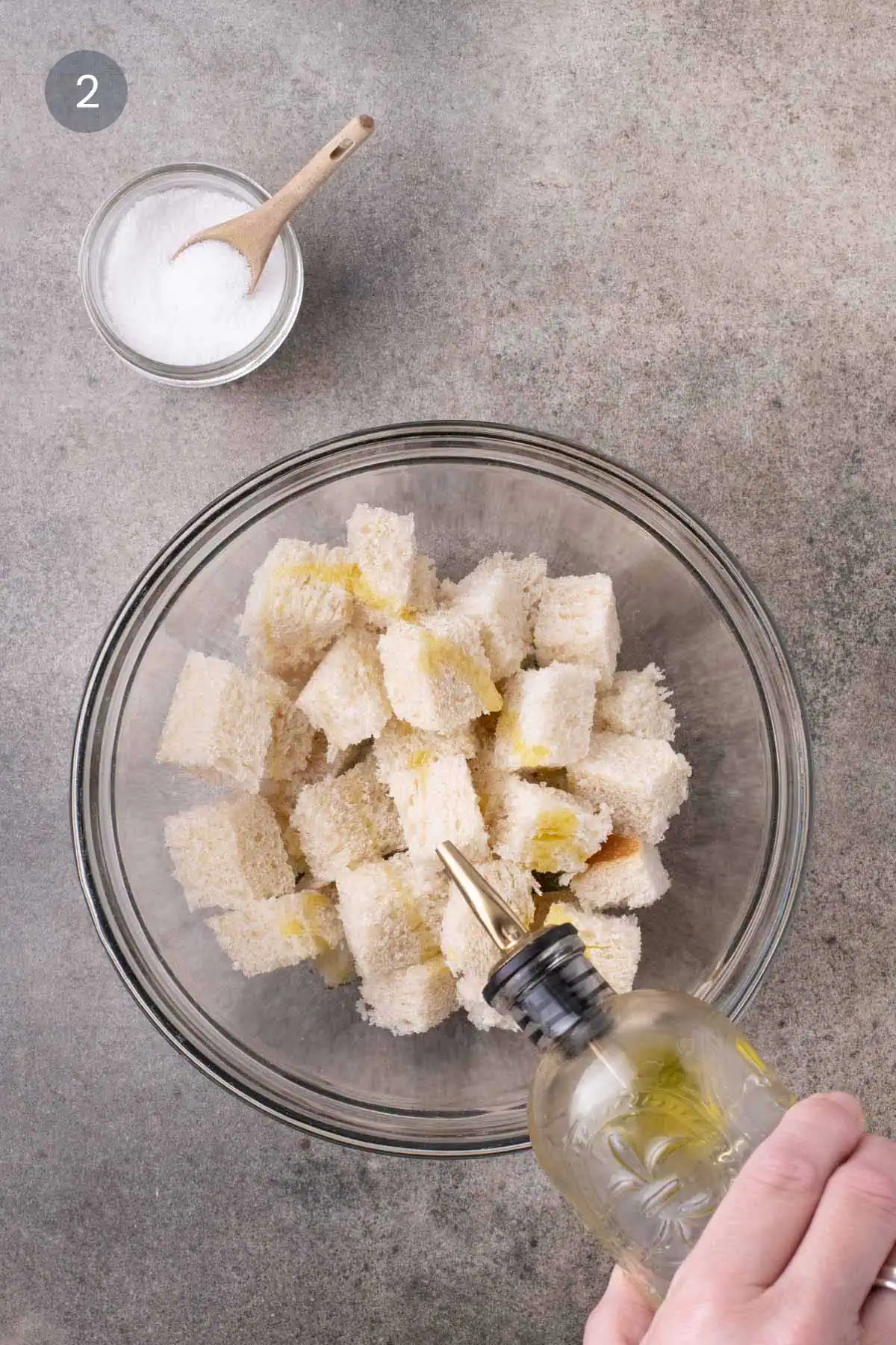 Olive oil being drizzled over bread cubes in a glass bowl, with a small bowl of salt nearby.