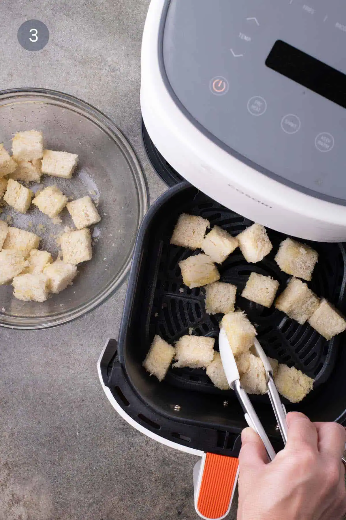 Silicone-tipped tongs placing bread cubes in a single layer in an air-fryer basket.