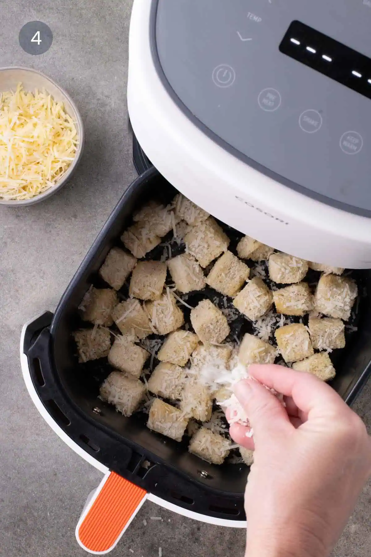 A hand sprinkling grated cheese over bread cubes in an air-fryer basket, with a bowl of cheese alongside.