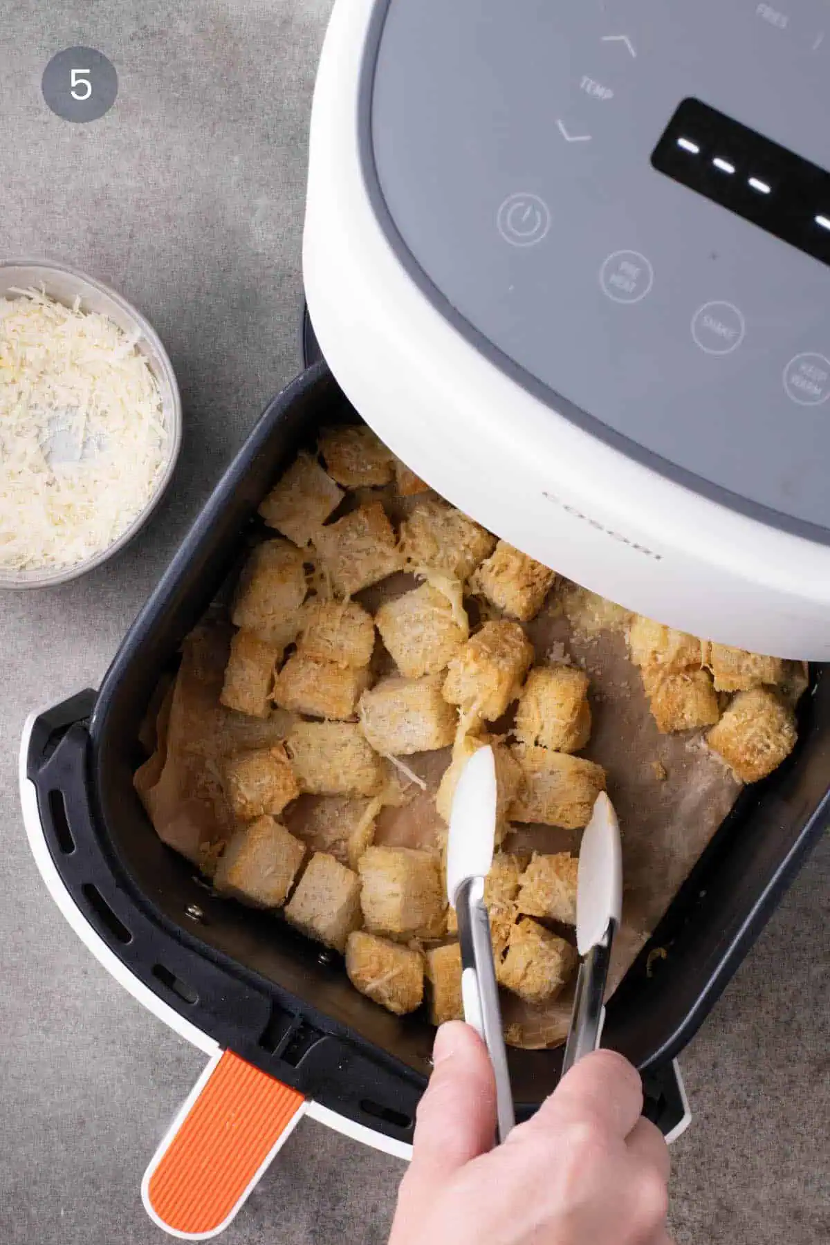 Silicone-tipped tongs turning lightly golden, cheese-coated croutons in a parchment-lined air-fryer basket.
