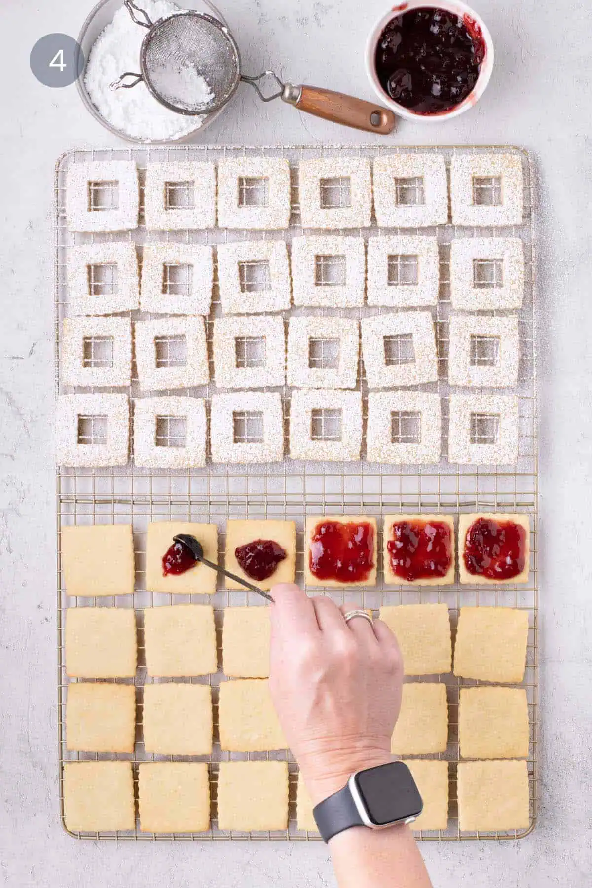 Hand spreading raspberry jam over baked fluted square cookie bases on a wire rack.