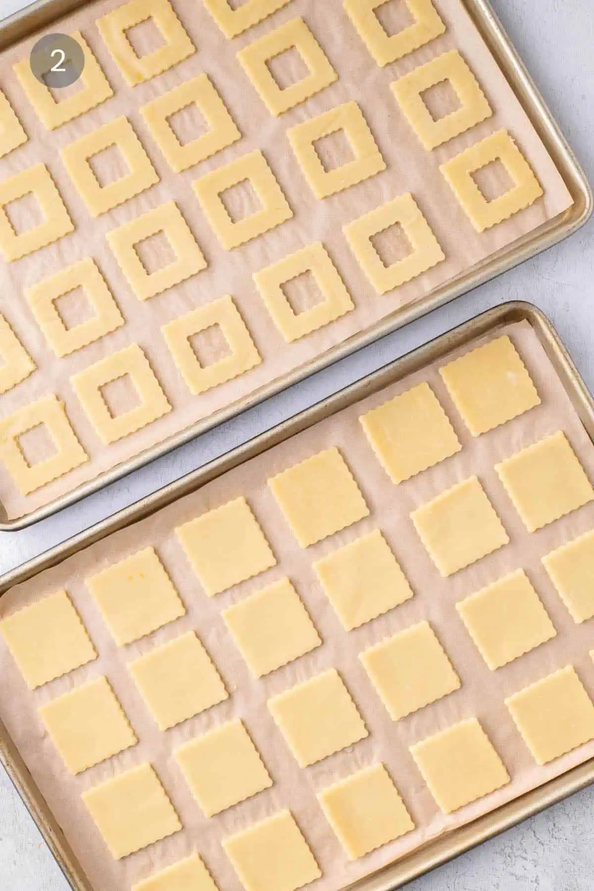 Unbaked fluted window tops and solid cookie bases arranged on two parchment-lined baking sheets.