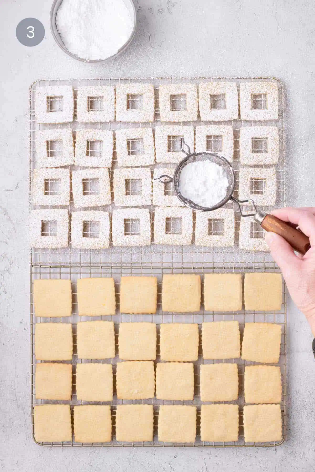 Powdered sugar being sifted over baked fluted window cookies on wire racks.