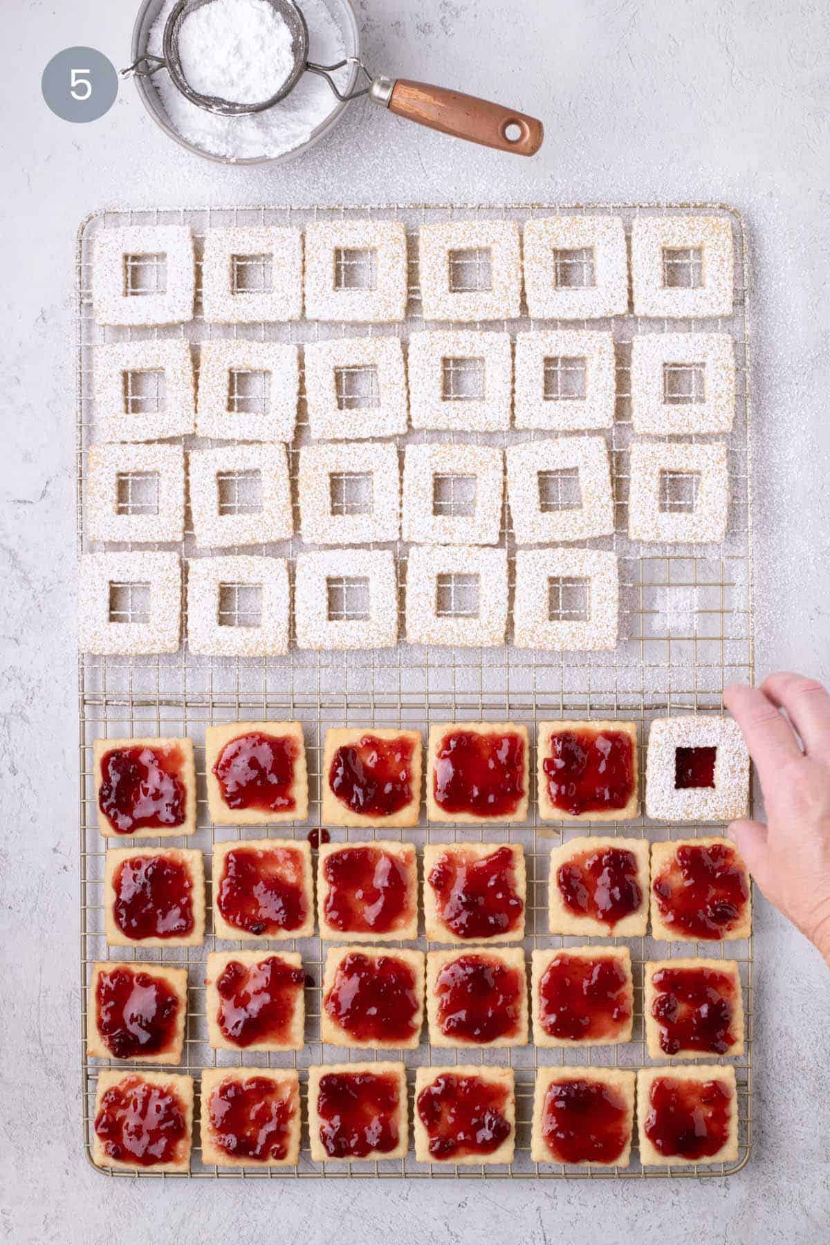 Hand placing a powdered-sugar-dusted window top over a raspberry-jam-covered cookie base.