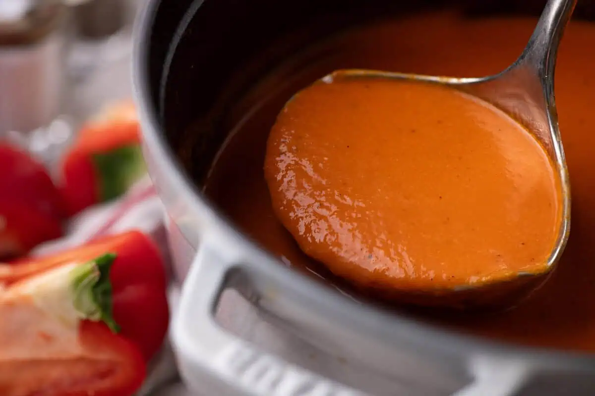 A ladle filled with smooth orange-red roasted pepper soup inside a Dutch oven, with red bell peppers in the background.