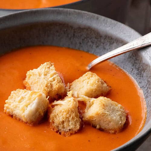 A gray ceramic bowl filled with smooth, orange-red roasted pepper soup, topped with golden cheese croutons and a silver spoon, with a pot of soup in the background.