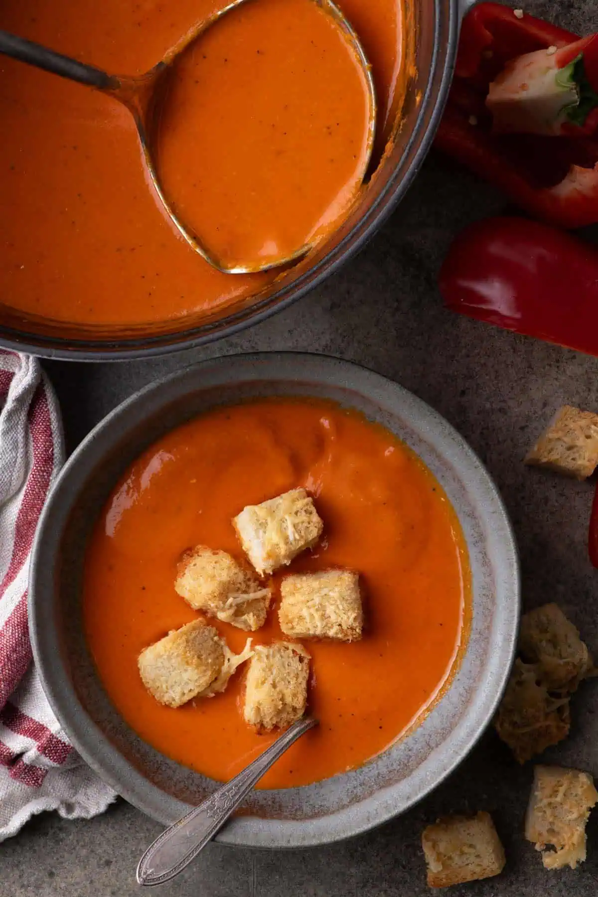 A Dutch oven filled with orange-red soup and a ladle, surrounded by red bell peppers and a plate of cheese croutons.