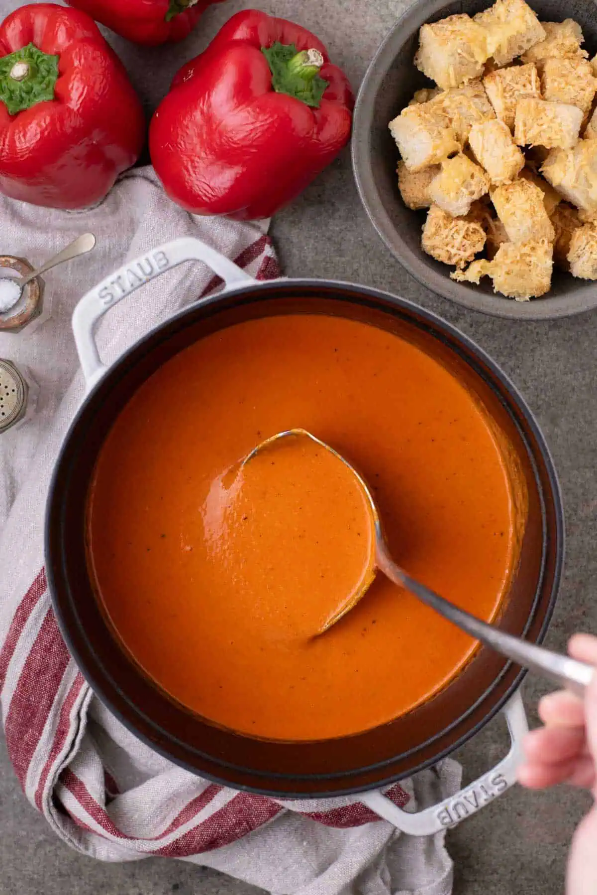 Overhead view of roasted red pepper soup in a pot and a serving bowl topped with cheese croutons, alongside red bell peppers.