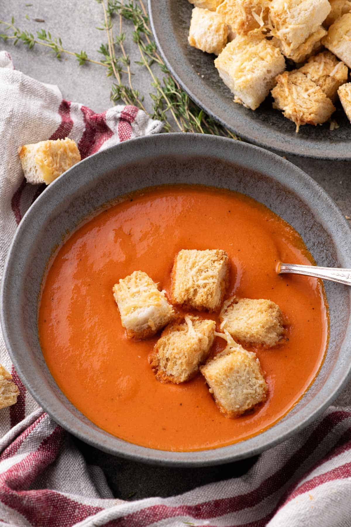 Overhead view of a Dutch oven filled with smooth orange-red soup, surrounded by fresh red peppers and a plate of cheese croutons.
