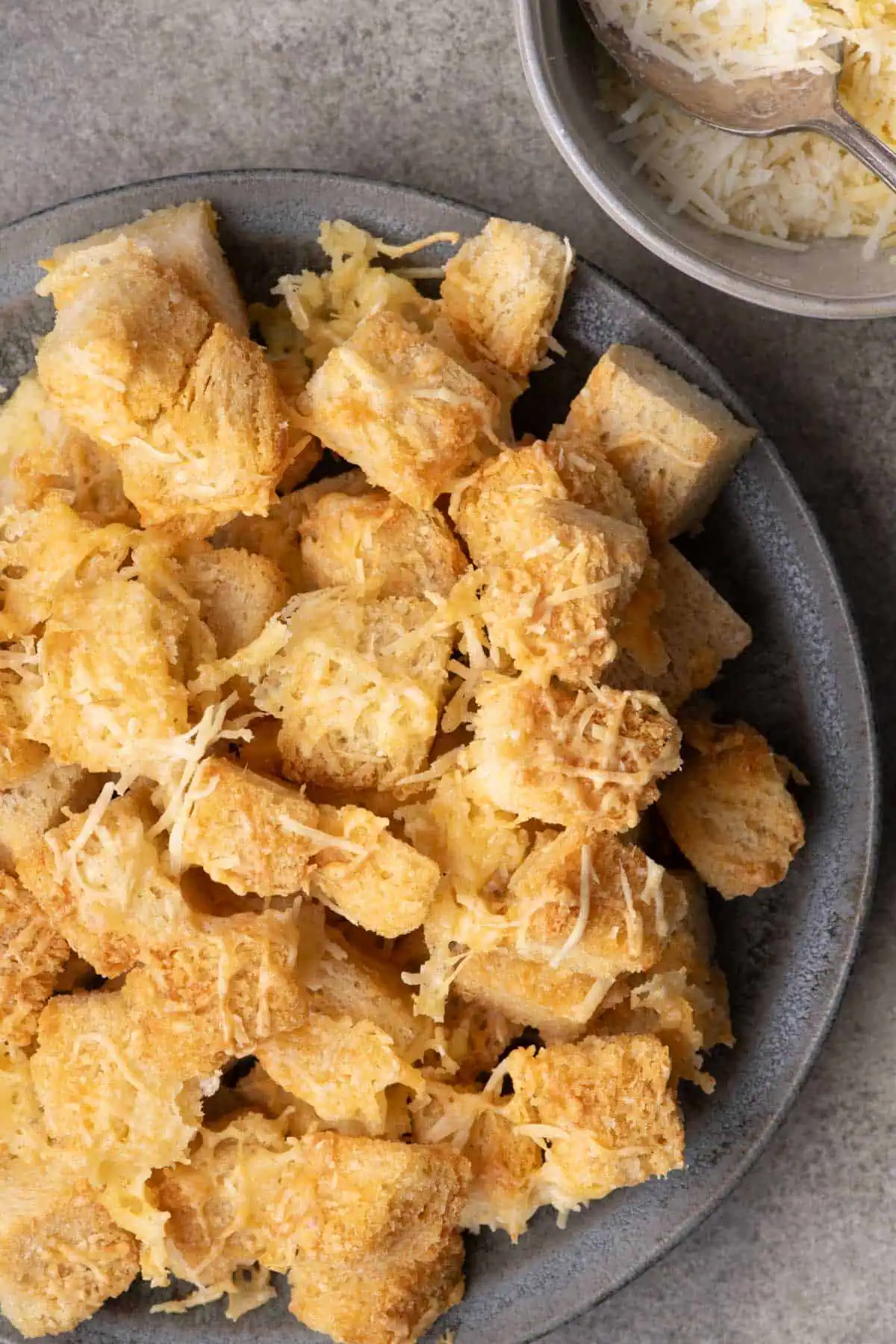 Golden air fryer croutons with melted cheese piled on a gray plate beside a bowl of grated cheese.