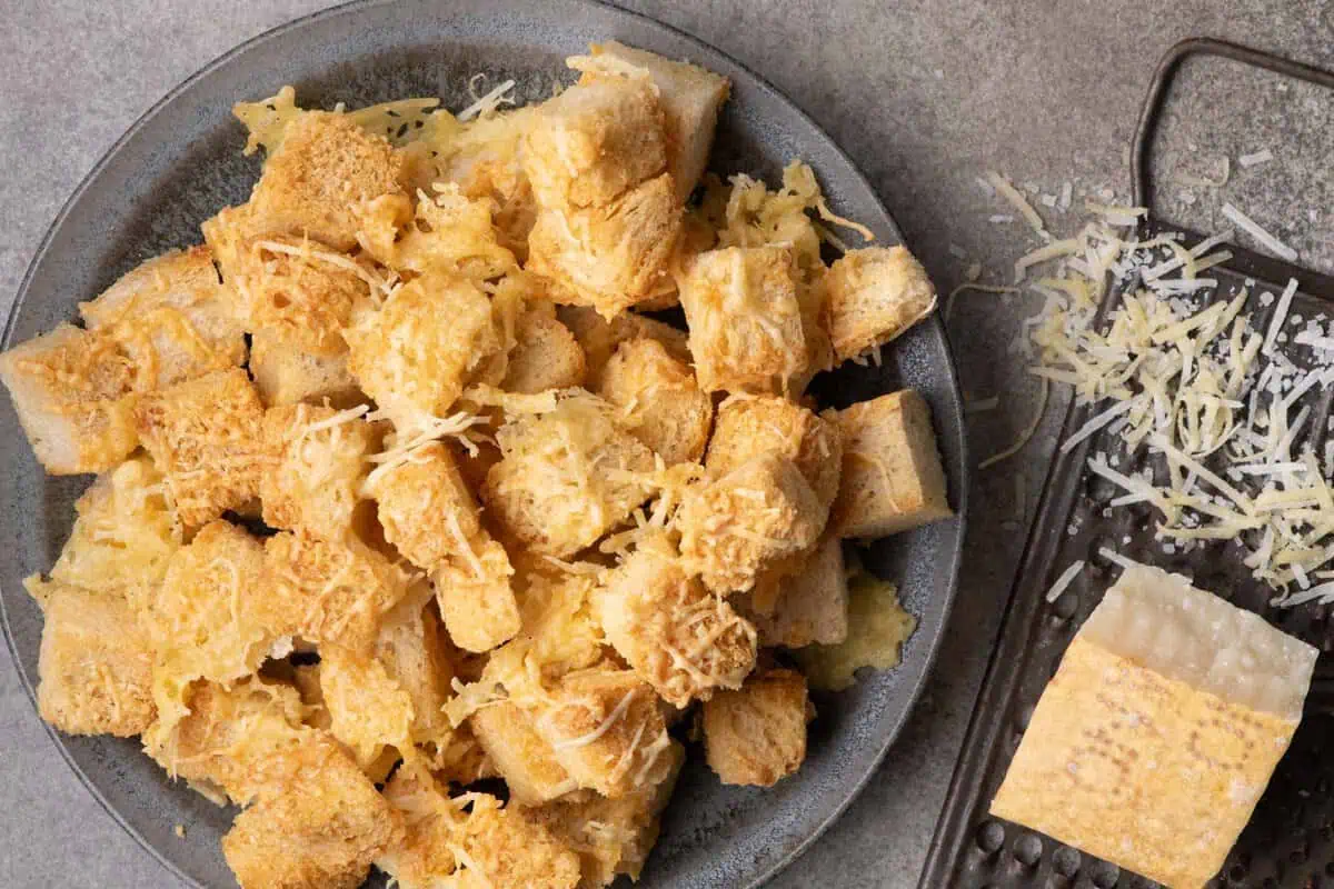 Overhead view of cheesy sourdough croutons on a gray plate beside a Parmesan wedge and grater.