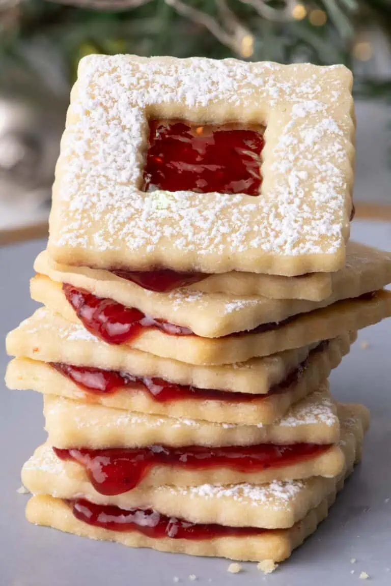 Stack of raspberry jam sandwich cookies dusted with powdered sugar in front of blurred evergreen branches and holiday lights.