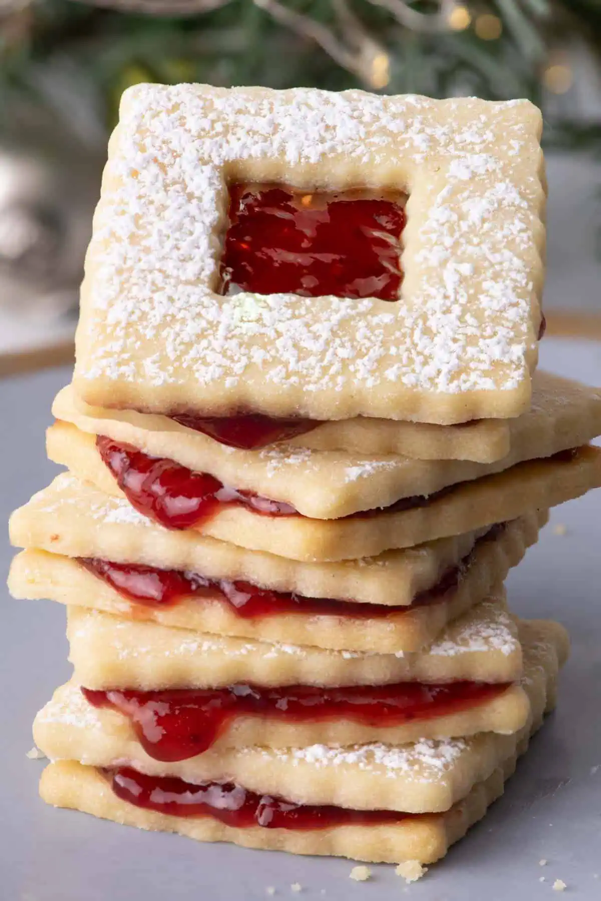 Stack of raspberry jam sandwich cookies dusted with powdered sugar in front of blurred evergreen branches and holiday lights.