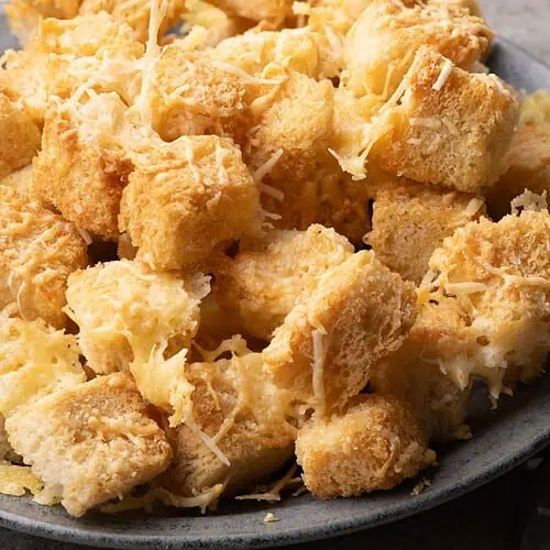 Close-up of golden sourdough croutons with melted cheese and crisp edges beside a Parmesan wedge.