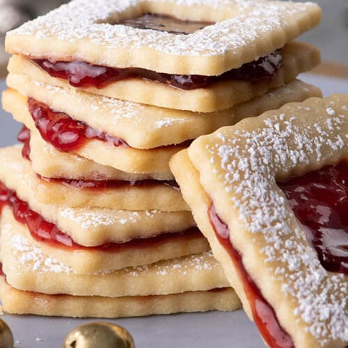 Raspberry jam sandwich cookies dusted with powdered sugar beside a gold jingle bell and blurred holiday greenery.