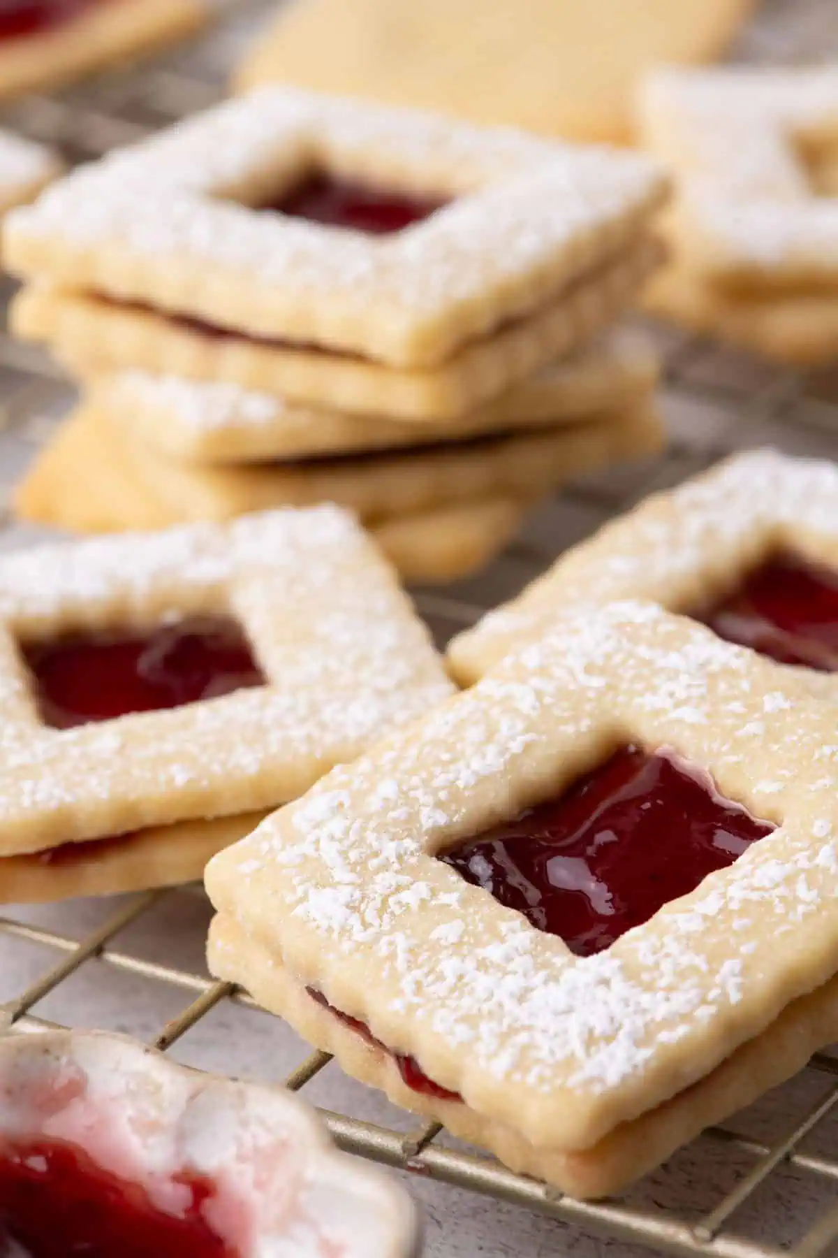 Raspberry jam sandwich cookies dusted with powdered sugar on a cooling rack with a bowl of raspberry jam.