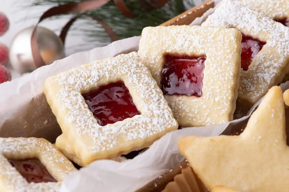 Square raspberry jam sandwich cookies dusted with powdered sugar beside star-shaped butter cookie.