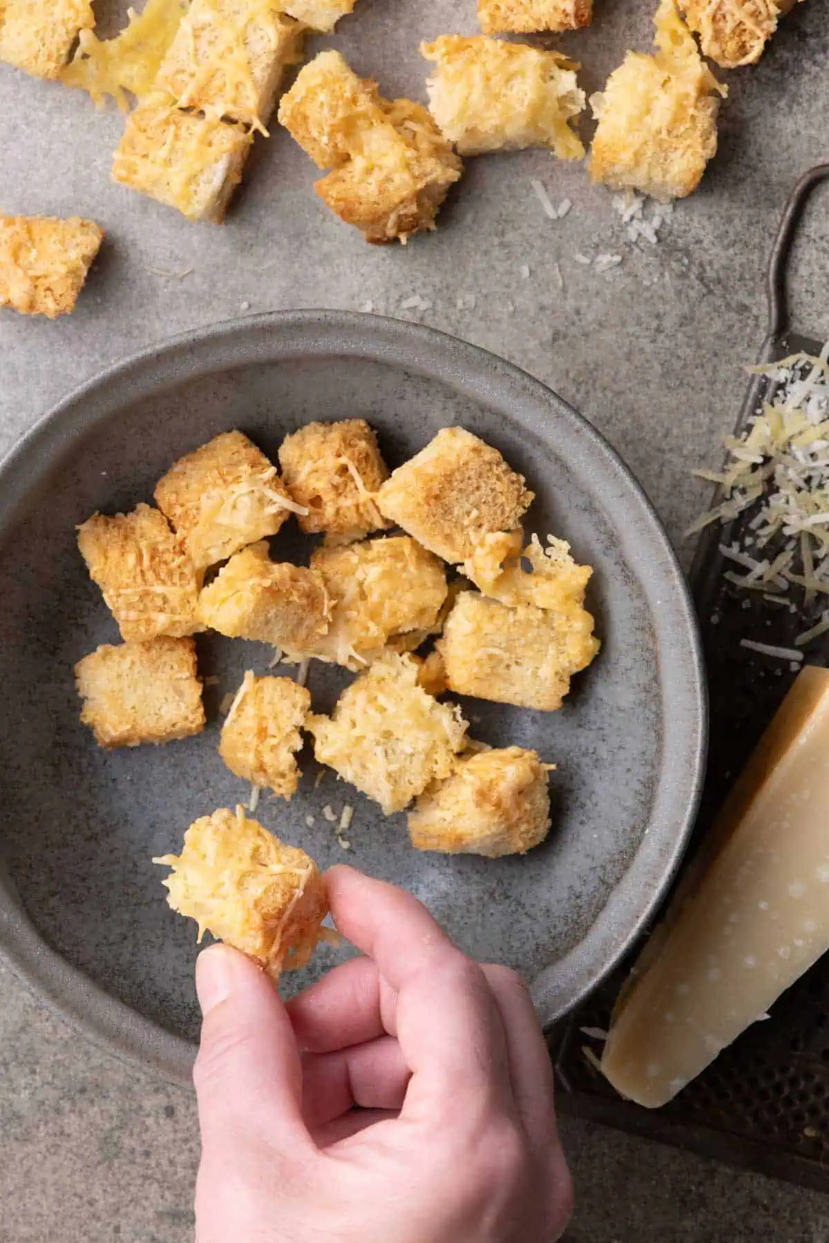 Hand holding a golden cheese-coated crouton above a gray plate beside a Parmesan wedge and grater.