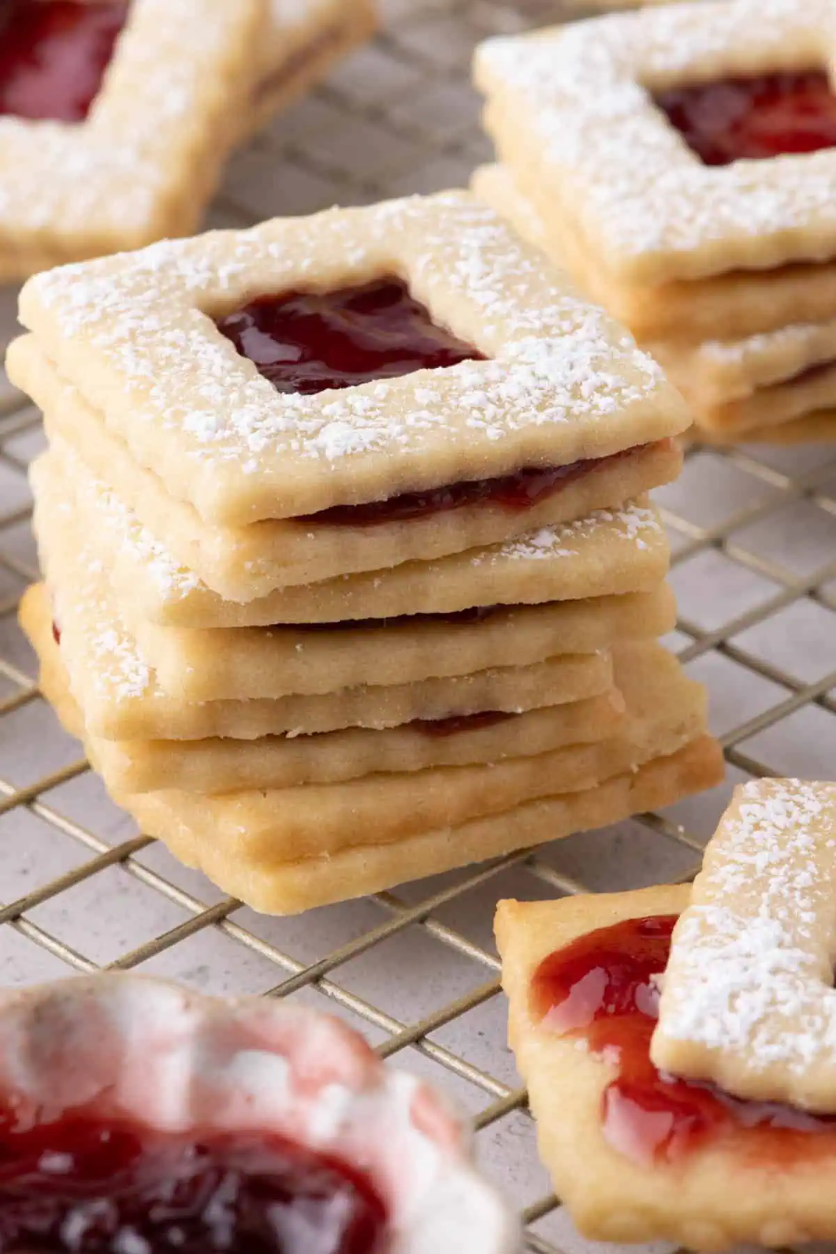 Stack of raspberry jam sandwich cookies dusted with powdered sugar on a cooling rack with a bowl of raspberry jam.