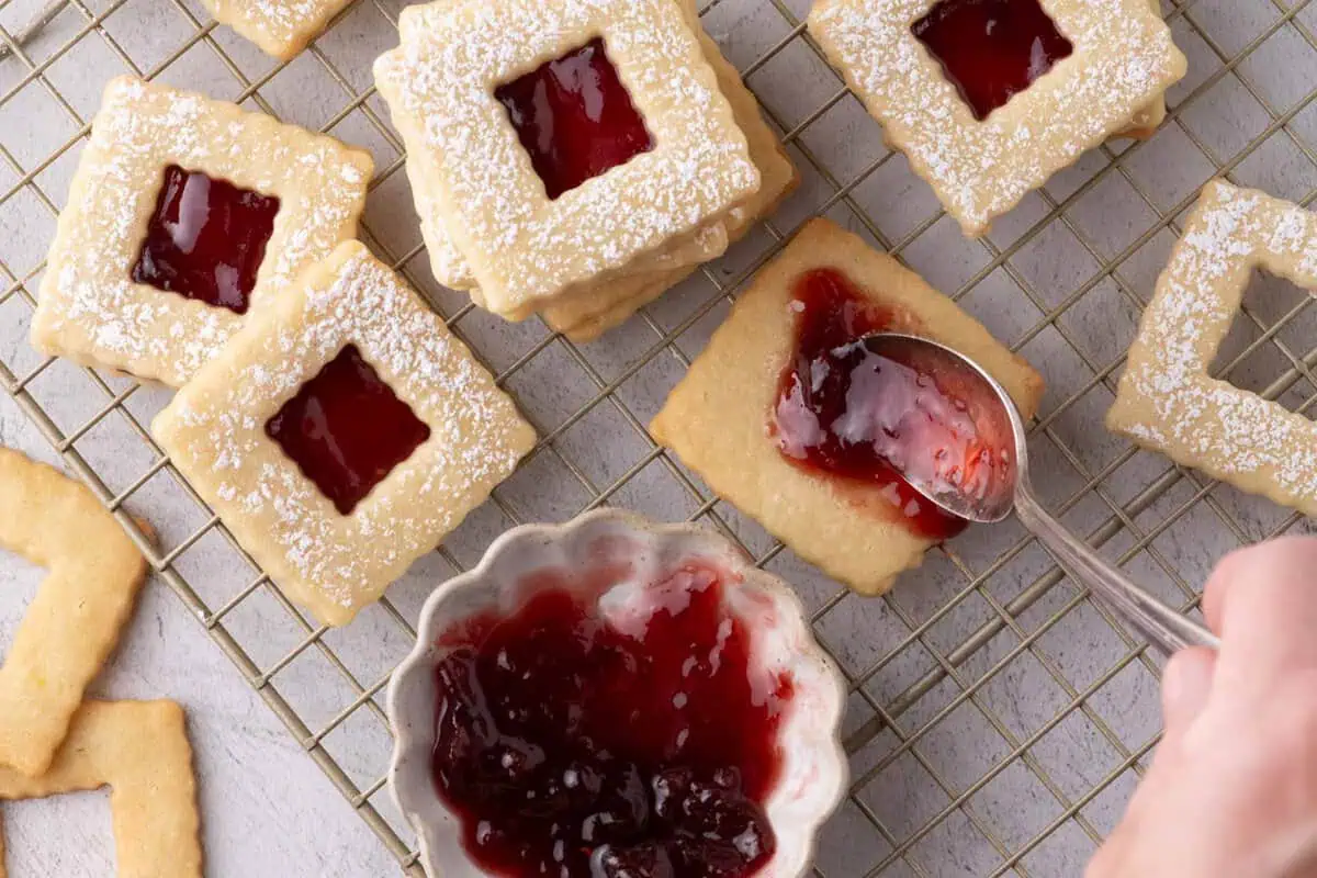Hand spreading raspberry jam on a square cookie beside powdered-sugar-dusted raspberry jam sandwich cookies on a cooling rack.