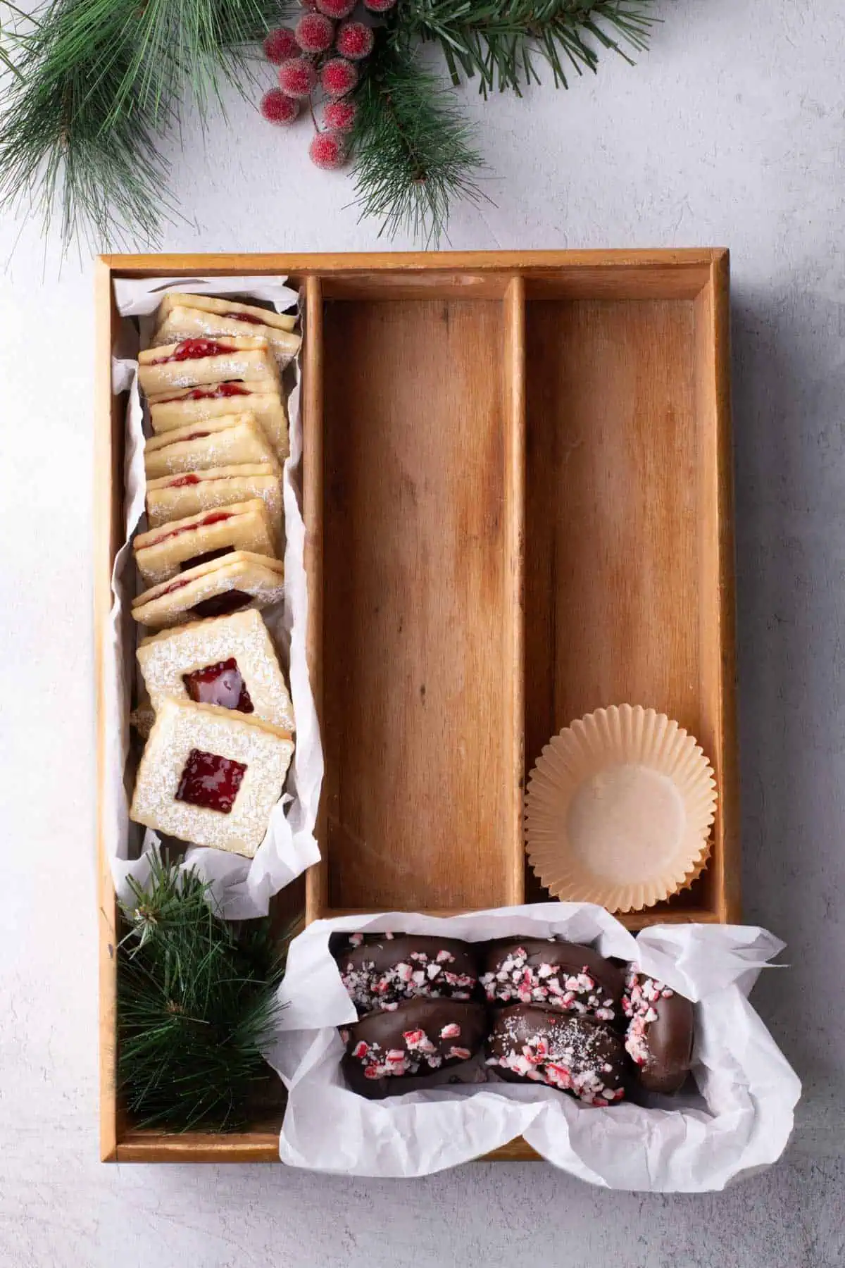 Partially filled Christmas cookie box with raspberry jam sandwich cookies and chocolate-dipped cookies arranged in a parchment-lined wooden tray