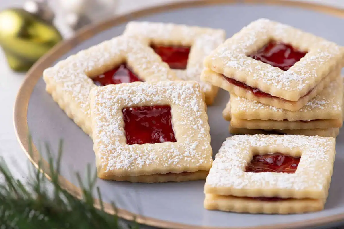 Raspberry jam sandwich cookies dusted with powdered sugar on a plate with a gold ornament and evergreen sprigs.