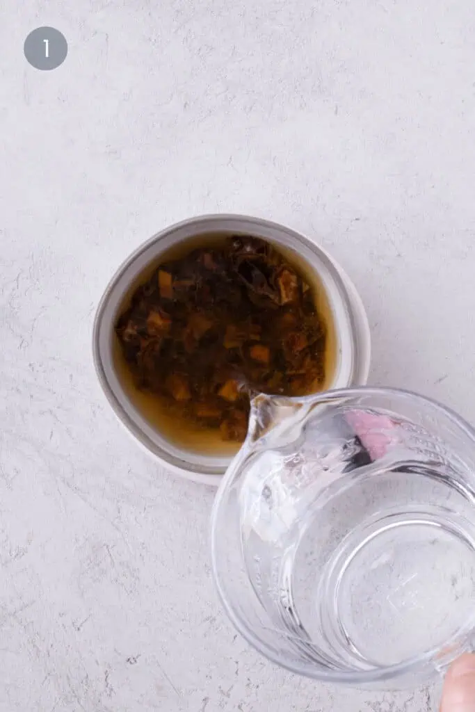 Water being poured from a glass measuring cup over diced dates in a gray bowl.