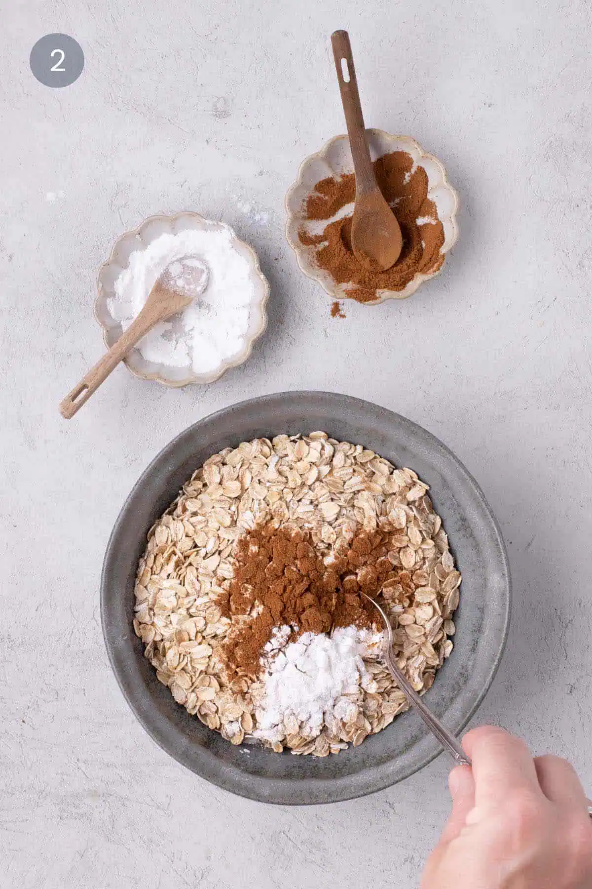 Rolled oats topped with cinnamon and baking powder in a gray bowl, with a measuring spoon held above.