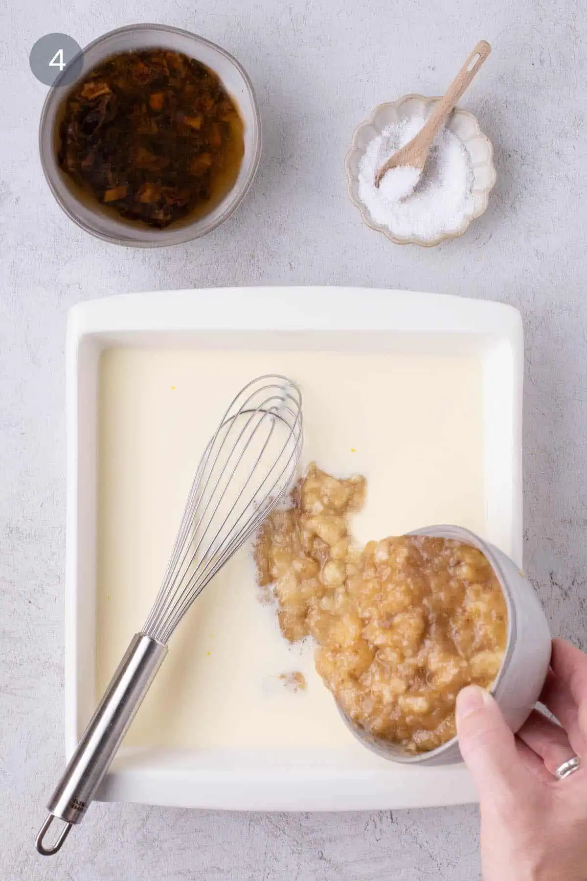 Mashed banana being added to the wet ingredients in a white baking dish, with a bowl of diced dates nearby.