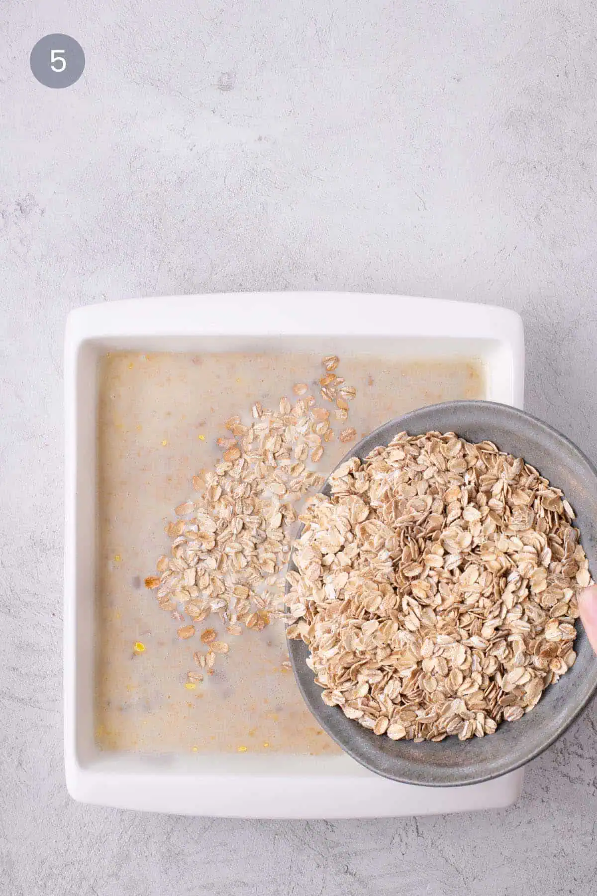 Rolled oats being tipped from a gray bowl into the wet banana oatmeal mixture in a white baking dish.