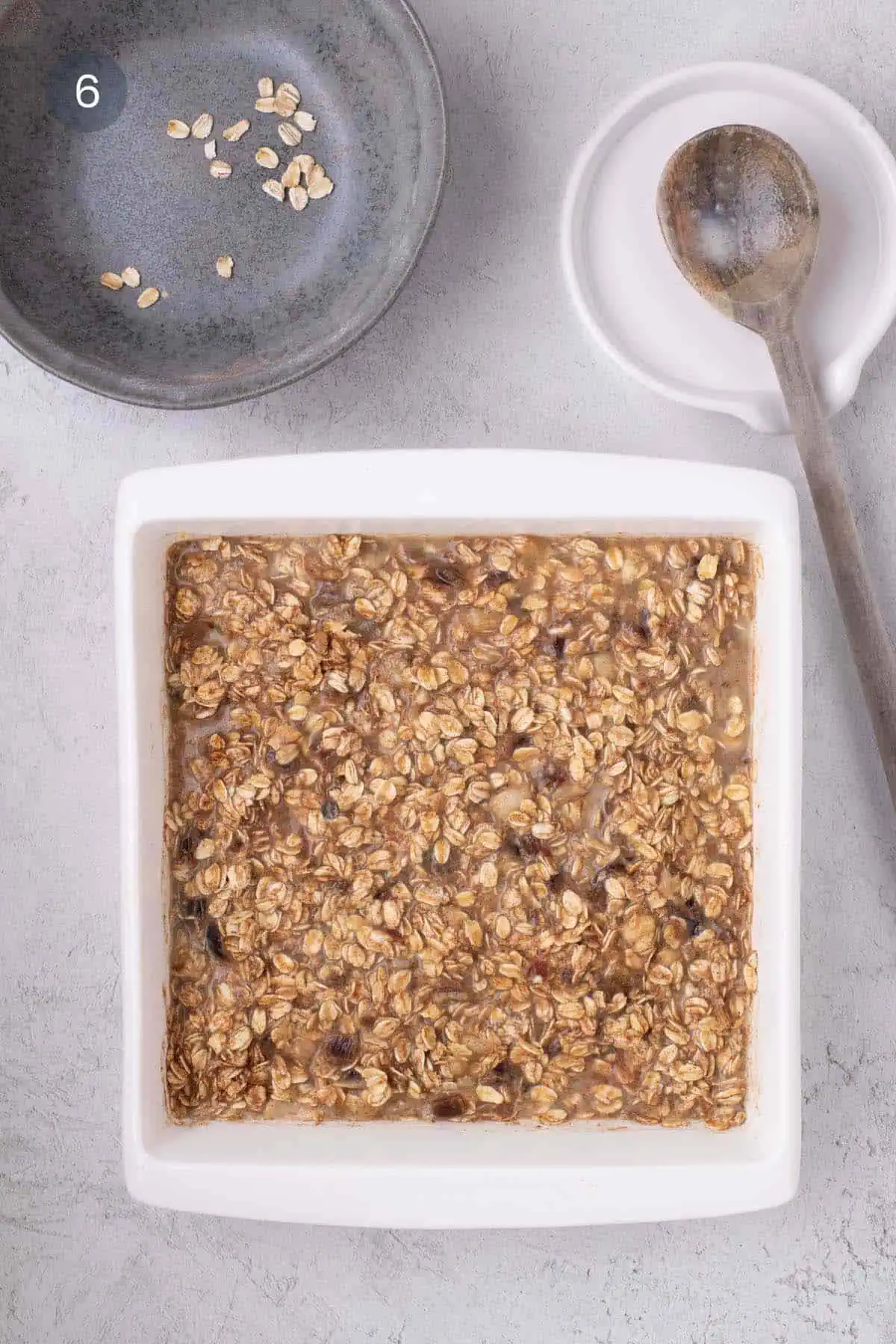 Unbaked oatmeal mixture with rolled oats and date pieces in a white square baking dish, with a wooden spoon beside it.